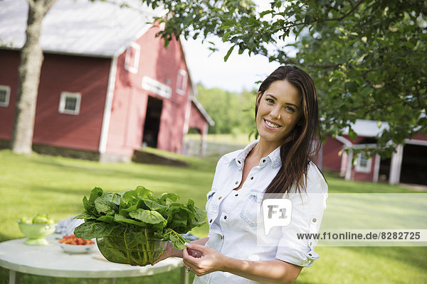 Eine Sommer-Familienzusammenkunft auf einem Bauernhof. Eine junge Frau mit langen braunen Haaren und hochgekrempelten Ärmeln trägt eine große Schüssel mit frischen grünen Salatblättern.
