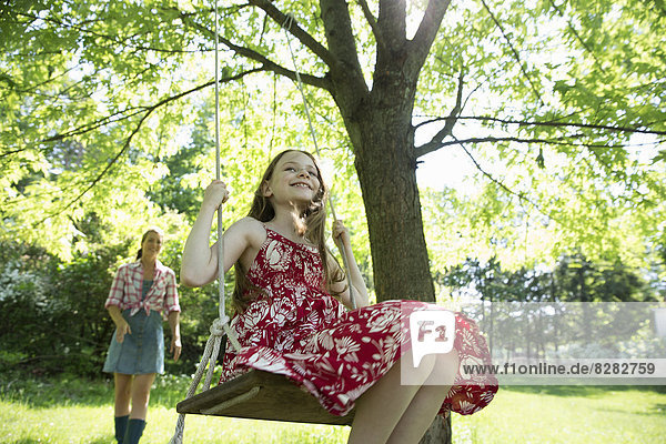 Sommer. Ein Mädchen in einem Sonnenkleid auf einer Schaukel  die an einem Baumzweig hängt. Eine Frau hinter ihr.
