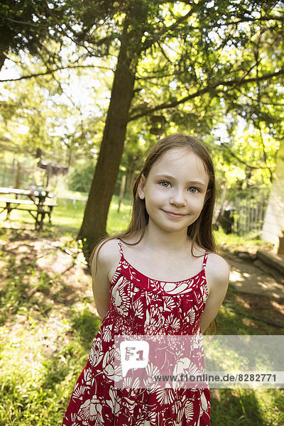 Ein junges Mädchen in einem roten  blumigen Sommerkleid im Schatten von Bäumen.