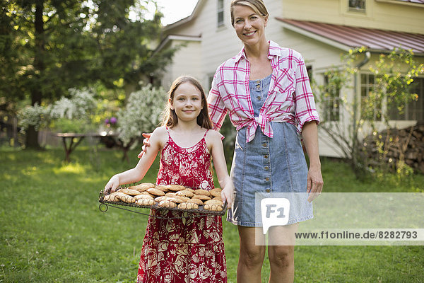 Hausgemachte Kekse backen. Ein junges Mädchen hält ein Tablett mit frisch gebackenen Keksen in der Hand  daneben eine erwachsene Frau.
