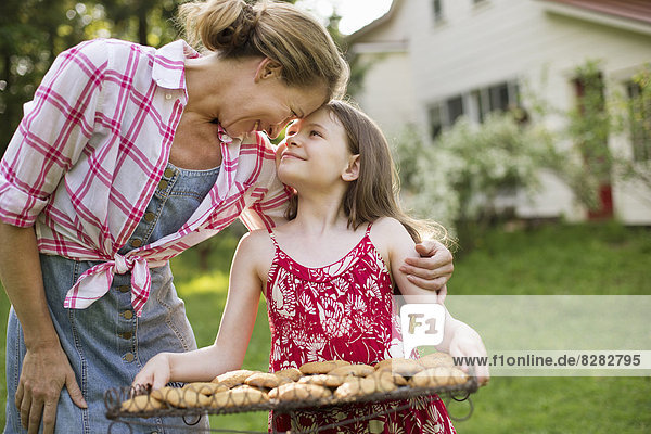 Hausgemachte Kekse backen. Ein junges Mädchen hält ein Tablett mit frisch gebackenen Keksen in der Hand  und eine erwachsene Frau beugt sich vor  um sie zu loben.