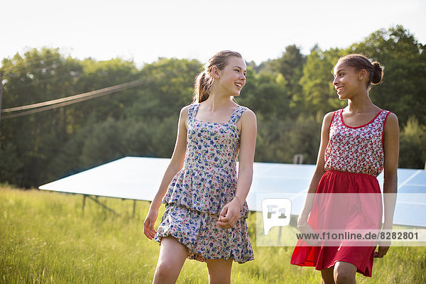 Zwei junge Mädchen auf dem Bauernhof  im Freien. Eine große Solaranlage auf dem Feld hinter ihnen.