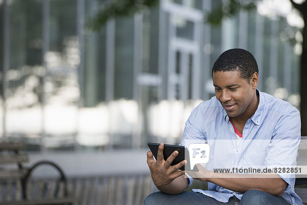 Sommer in der Stadt. Menschen im Freien  die unterwegs in Kontakt bleiben. Ein Mann sitzt auf einer Bank mit einem digitalen Tablet.