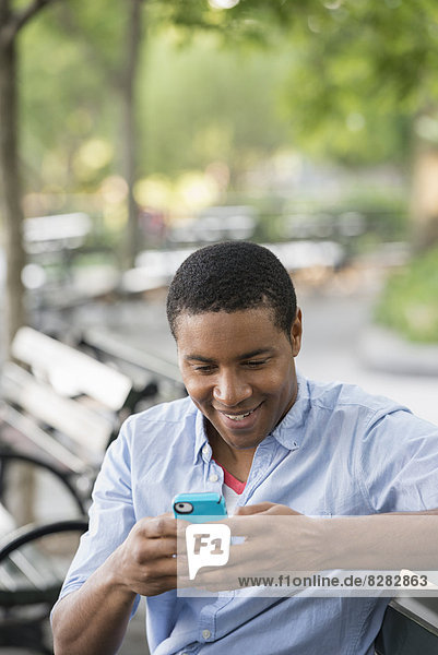 Sommer in der Stadt. Ein Mann sitzt auf einer Bank und benutzt ein Smartphone.