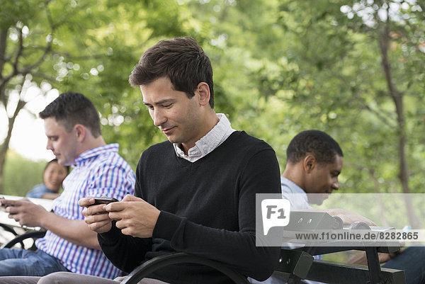 Sommer in der Stadt. Eine Frau und drei Männer sitzen im Park  jeder mit seinem eigenen Telefon oder mit einem Tablet.