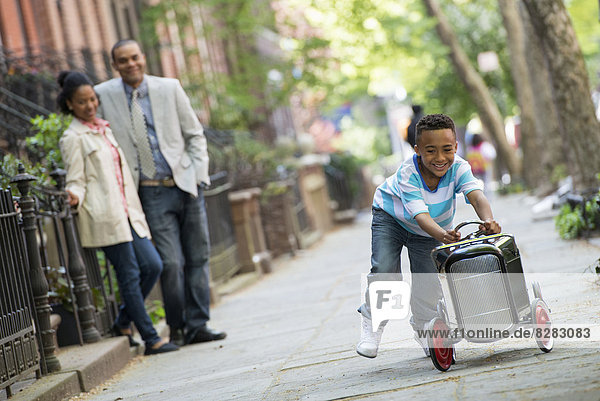 Ein junger Junge spielt mit einem altmodischen Spielzeugauto auf Rädern auf einer Straße der Stadt. Ein Pärchen schaut zu.