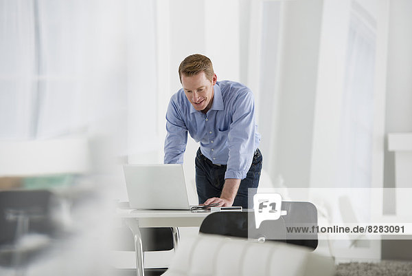 Wirtschaft. Ein Mann  der über einem Schreibtisch steht und sich herunterlehnt  um einen Laptop-Computer zu benutzen.