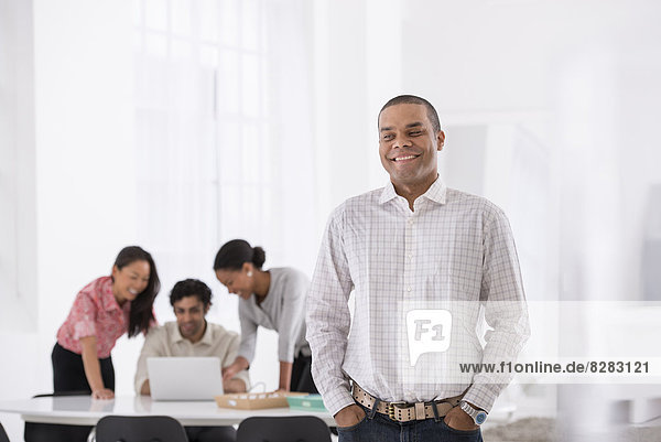 Business. A Man Standing With His Hands In His Pockets  And Three People Leaning Over A Desk Behind Him.