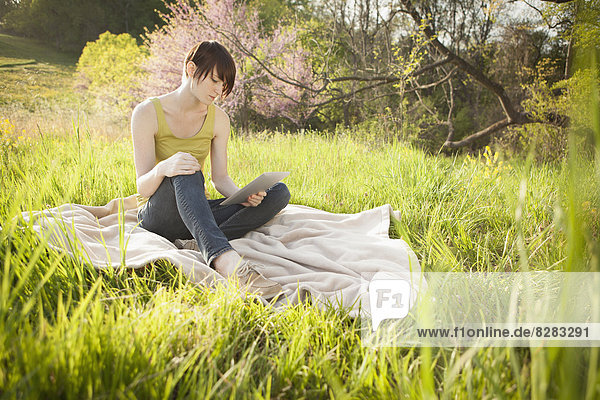 A Young Woman Sitting In A Field  On A Blanket  Reading From A Digital Tablet.