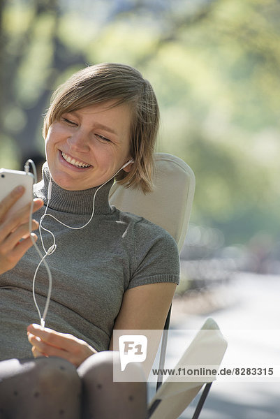 City Life. A Woman Sitting In A Camping Chair In The Park  Listening To Music Wearing Headphones.