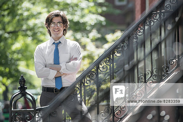 City. A Young Man In W White Shirt And Blue Tie  Standing With Arms Folded Outside A Townhouse  On The Steps.