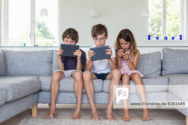 Geschwister auf Sofa mit digitalen Tabletts und Handy
