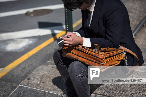 Businessman sitting on kerb with satchel  smoking cigarette
