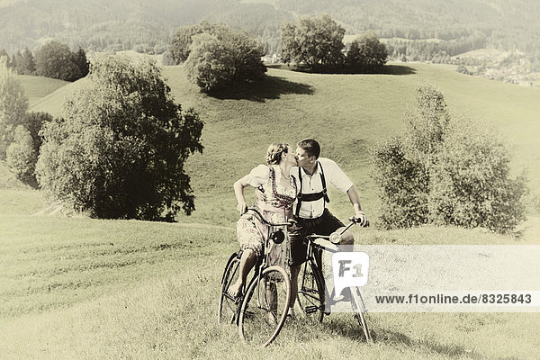 Man wearing leather pants and a woman wearing a dirndl kissing on old bicycles within a natural landscape