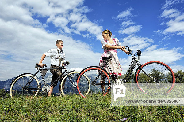 Man wearing leather pants and a woman wearing a dirndl with old bicycles within a natural landscape