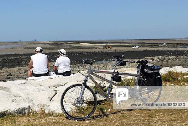 'Bike ride on the ''île Madame'' island: couple near their tandem looking at the sea at low tide with Fort Boyard far off.'
