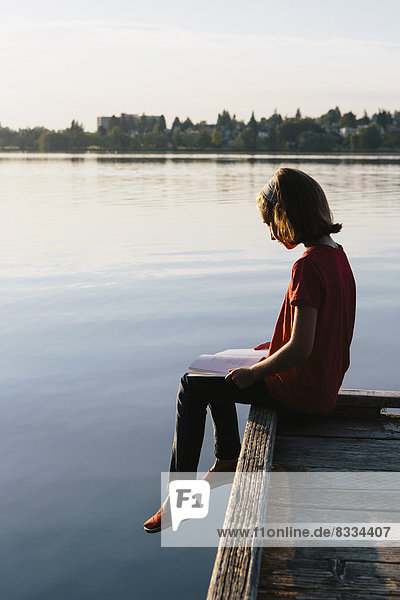 A young girl sitting on a dock  reading a book.