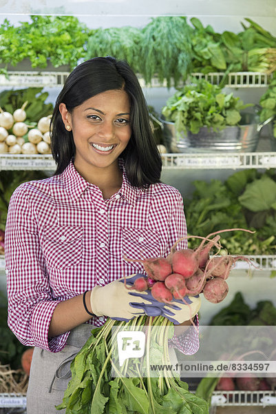 Ein Bauernstand mit Reihen von frisch gepflücktem Gemüse zum Verkauf. Eine Frau hält ein Bündel Karotten in der Hand.