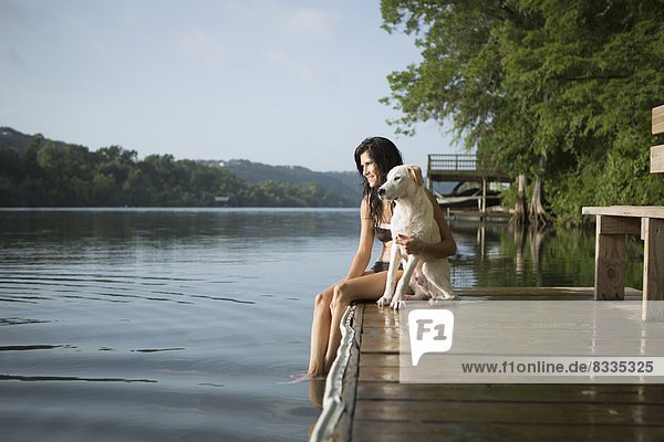 Eine Frau mit dem Arm um einen kleinen weißen Hund auf einem Steg an einem See.