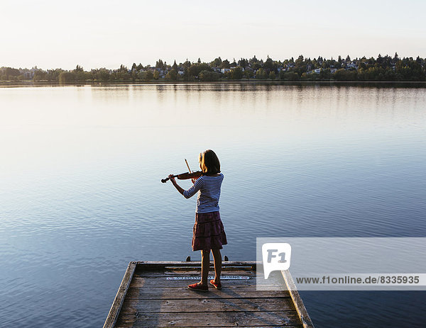 Ein zehnjähriges Mädchen spielt im Morgengrauen auf einem hölzernen Steg Geige.