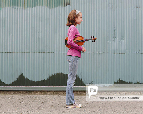 Ein zehnjähriges Mädchen,  das eine Geige unter dem Arm hält und auf einer Straße steht.