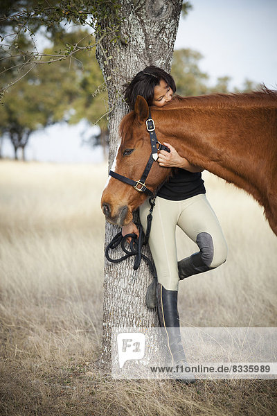 Eine Frau steht unter einem Baum auf einem Feld  hält ein Pferd am Halfter und streichelt seinen Kopf.