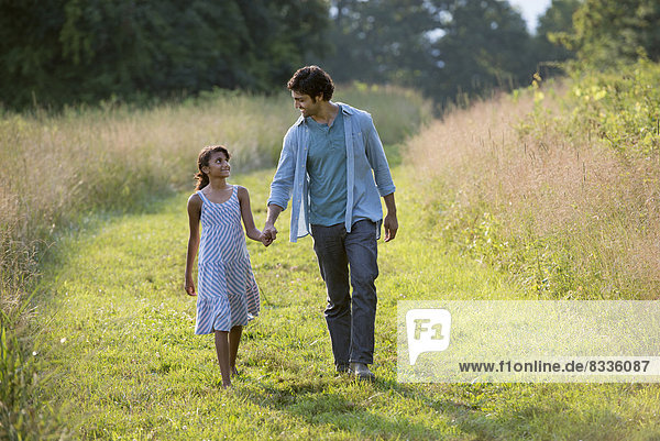 A man and a young girl walking down a mown path in the long grass holding hands.