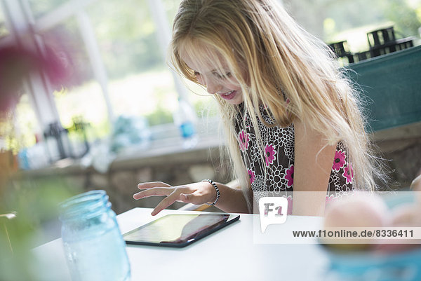A young girl in a flowered dress using a digital tablet. Sitting at a table.