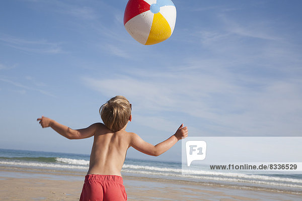 Ein Junge in Badehose am Strand  über ihm ein großer Wasserball in der Luft.