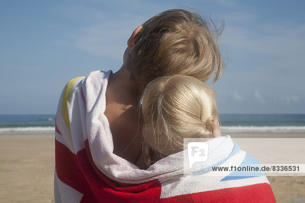 Zwei Kinder teilen sich ein Handtuch. Rückenansicht eines Bruders und einer Schwester  die am Strand sitzen und aufs Meer blicken.