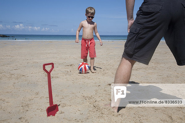 Ein Junge spielt mit einem Mann auf dem Sand Fußball. Vater und Sohn. Ein Strandspaten  der aufrecht im Sand steht.
