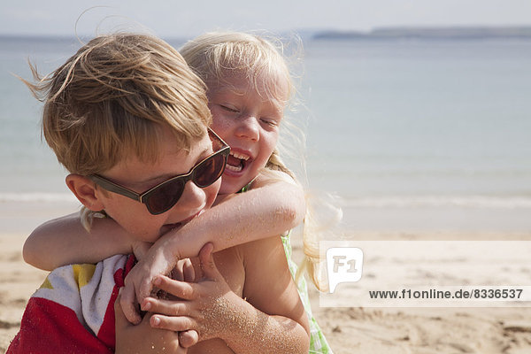 A brother and sister play fighting on the beach. A boy in sunglasses and a younger girl with her arms around his neck.