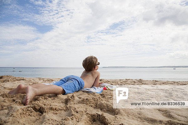 A boy lying on his front on the sand looking out to sea.