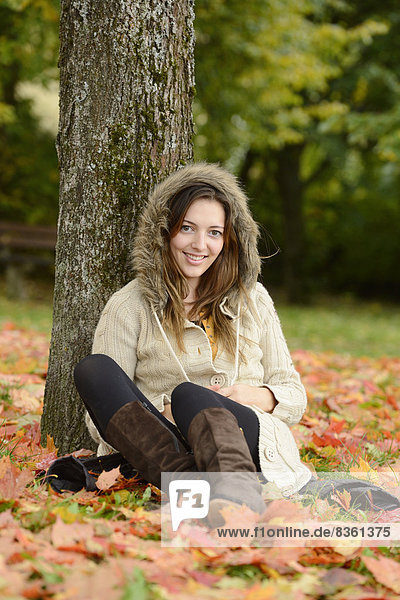 Lächelnde junge Frau sitzt unter einem Baum im Herbst