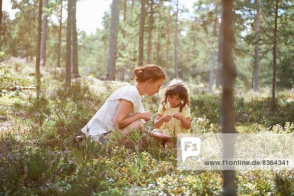 Mutter und Tochter sitzen im langen Gras und schauen auf Pflanzen.