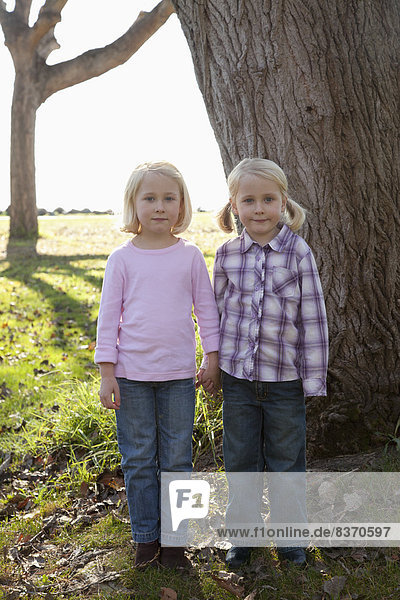 Two Young Girls With Blond Hair Hold Hands Standing In Front Of A Large Tree Trunk Crab Cove  California  United States Of America