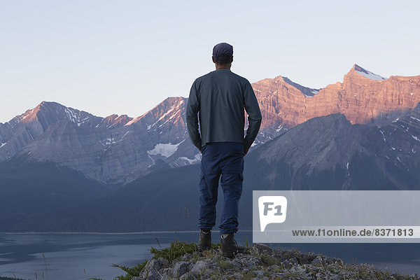 A Man Stands On A Ridge Overlooking A Lake Towards The Rocky Mountain Peaks Kananaskis  Alberta  Canada