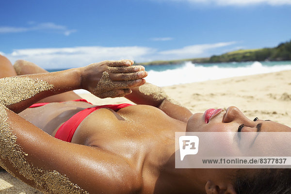 A young woman in a red bikini lays on a sandy beach at the water's edge Maui  Hawaii  United States of America