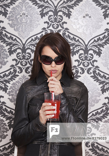 Junge Frau mit Sonnenbrille und rotem Getränk vor dekorativer Tapete