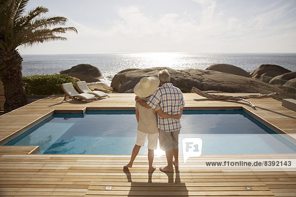 Seniorenpaar umarmt vom modernen Pool mit Blick aufs Meer