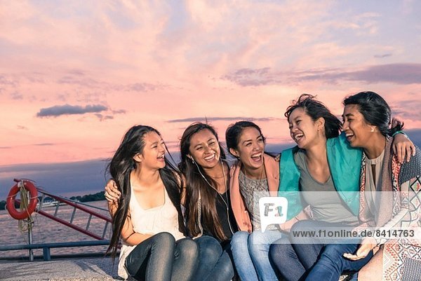 Young women lying sitting at waterfront at dusk with arms around
