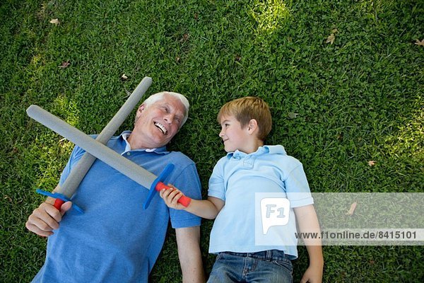 Großvater und Enkel auf Gras liegend mit Spielzeugschwertern