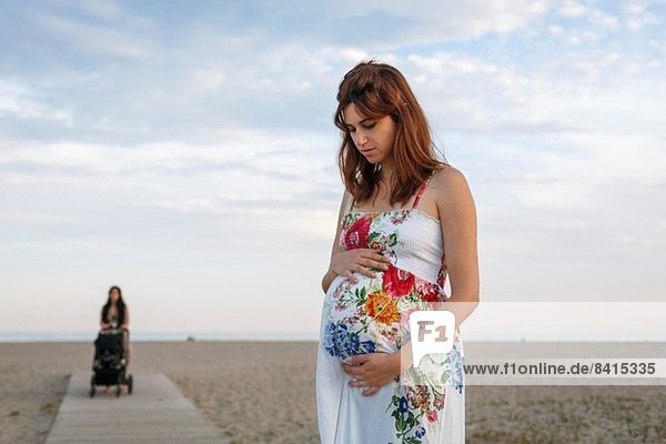 Schwangere Frau steht auf der Strandpromenade  Frau schiebt Kinderwagen im Hintergrund