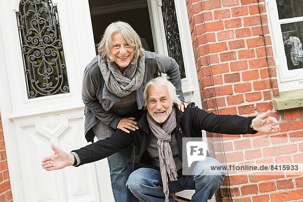 Älteres Ehepaar vor der Haustür  Mann kauert mit offenen Armen