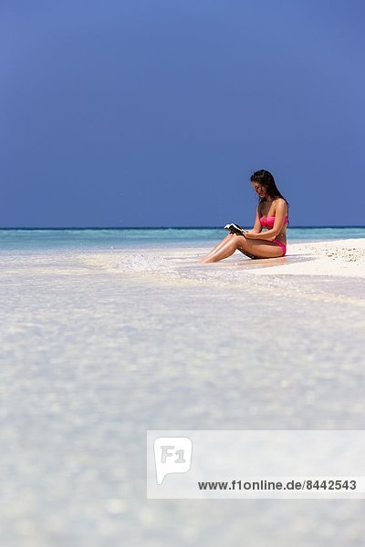Maldives  Young woman in bikini sitting in shallow water reading book