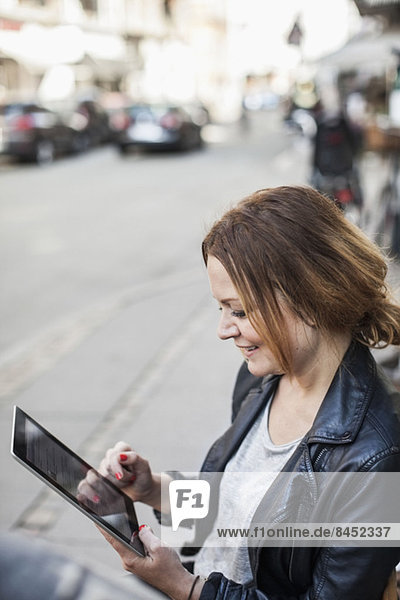 Lächelnde mittlere erwachsene Frau mit digitalem Tablett auf der Bürgersteigbank