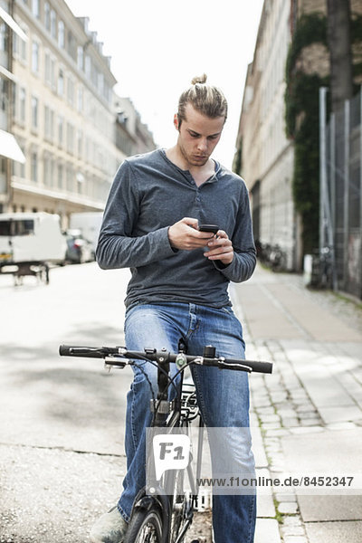 Junger Mann  der mit dem Handy auf dem Fahrrad auf der Straße sitzt