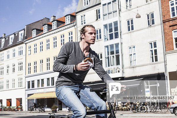 Junger Mann mit Burger beim Radfahren auf der Stadtstraße