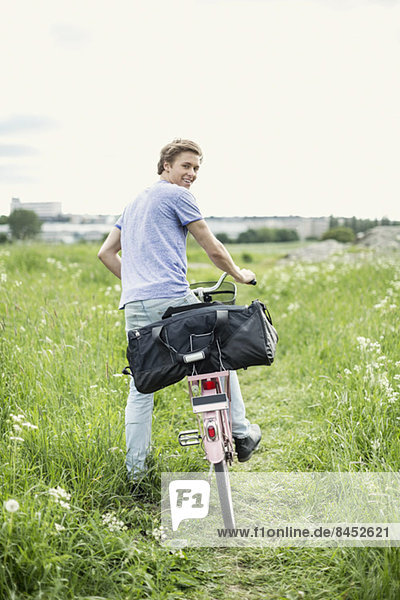 Rear view portrait of young man with bag and bicycle standing on field
