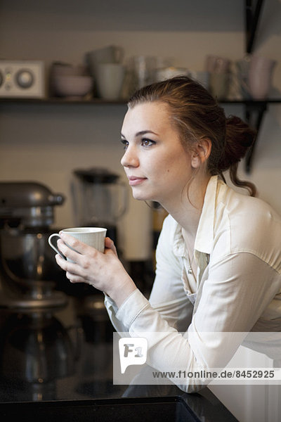 Nachdenkliche Frau mit Kaffeetasse schaut weg und lehnt sich an die Küchenzeile.